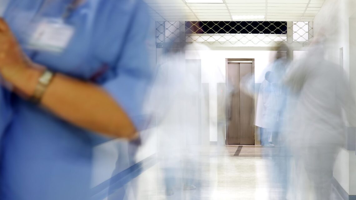 A blurred hospital corridor with medical staff in blue and white uniforms moving quickly, creating a sense of motion. A lift is visible at the end of the hallway.