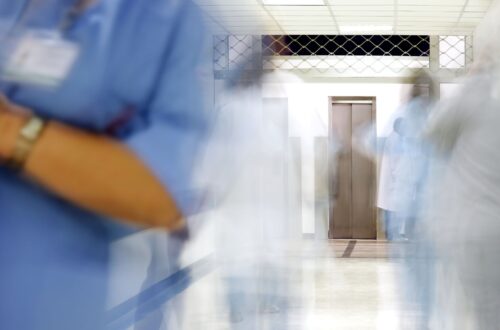 A blurred hospital corridor with medical staff in blue and white uniforms moving quickly, creating a sense of motion. A lift is visible at the end of the hallway.