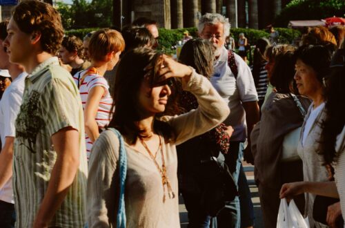 A busy street filled with people walking in different directions under strong sunlight, with one woman shielding her eyes from the glare.
