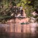 Two people paddling a canoe along a calm river beside trees and a stone building, symbolising community, connection and shared journeys.