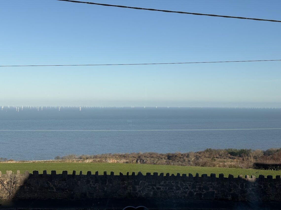 View of the sea under a bright blue sky, with white wind turbines on the horizon. In the foreground, green fields and a stone wall.”