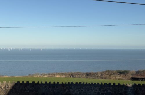 View of the sea under a bright blue sky, with white wind turbines on the horizon. In the foreground, green fields and a stone wall.”