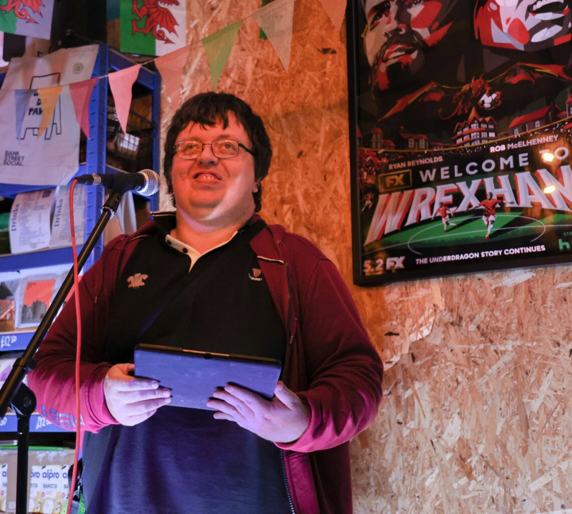 Poet performing at a VoiceBox Wrexham spoken-word open mic, standing at a microphone and reading from a Kindle.