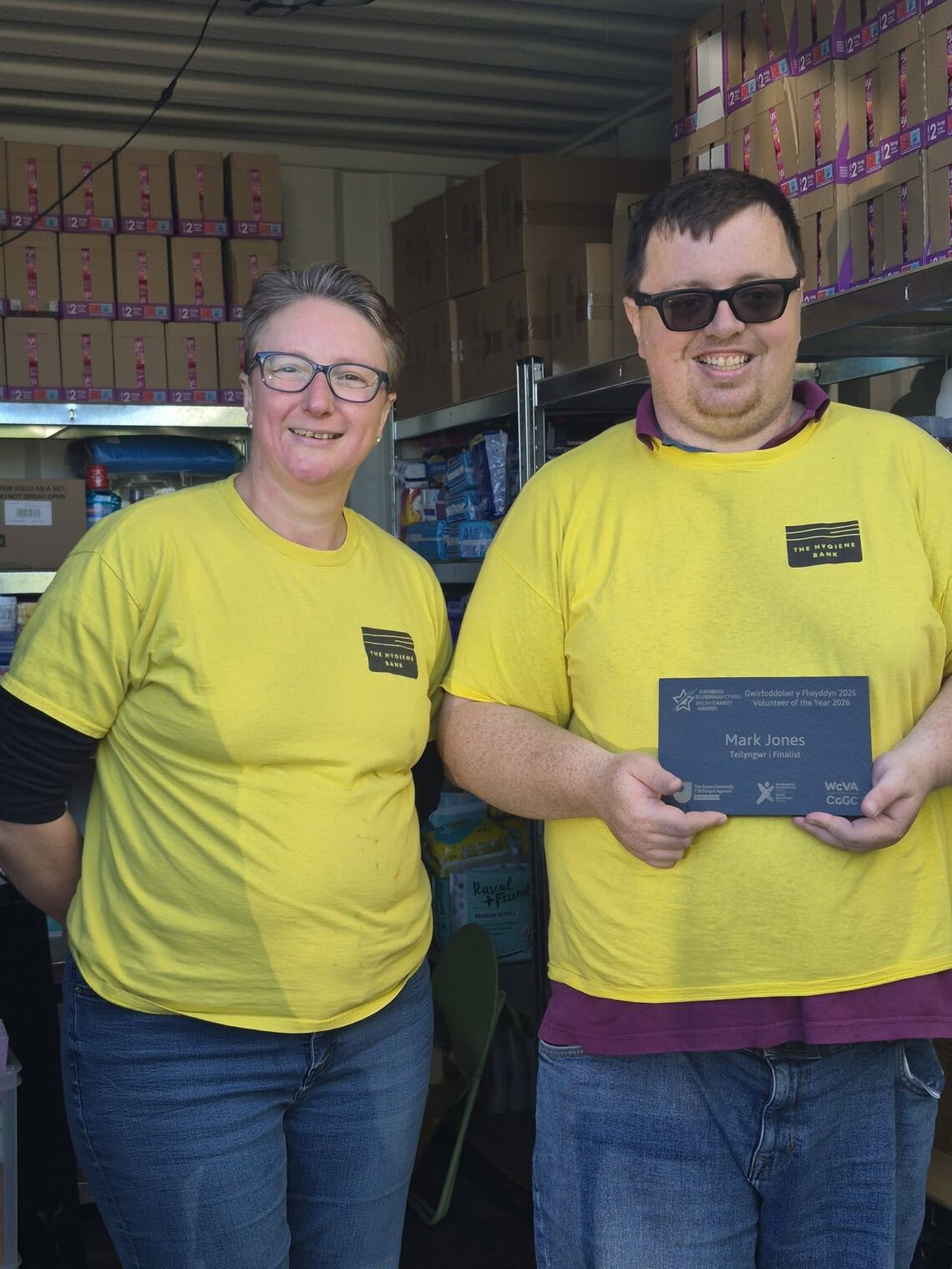 Mark Jones and Debbie Fretwell at The Hygiene Bank North Wales storage facility, standing beside shelves of donated hygiene products.